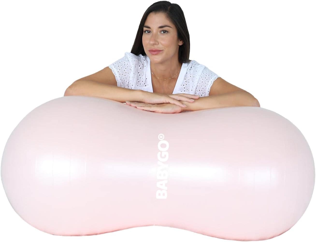 A person sits behind a large, light pink inflatable peanut-shaped exercise ball with BABYGO printed on it. Wearing a white blouse and resting their arms, they look forward, contemplating the ideal hospital bag items for labor induction.