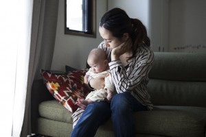 A woman sits on a couch holding a baby on her lap. The woman, possibly dealing with postpartum anxiety, looks down at the baby while resting her chin on her hand. They are indoors, near a window with light streaming in. A patterned pillow is behind them on the couch.