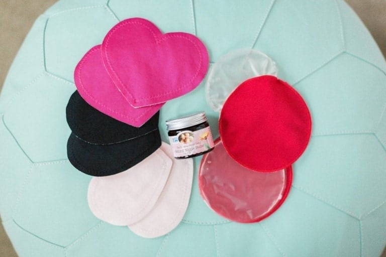 The image shows an assortment of reusable cloth pads in various shapes and colors, including heart-shaped and circular pads in pink, black, white, and translucent silicone. Ideal for those dealing with cracked nipples or engorgement, a small jar of "Earth Mama Organic Nipple Butter" is placed in the middle of the pads.