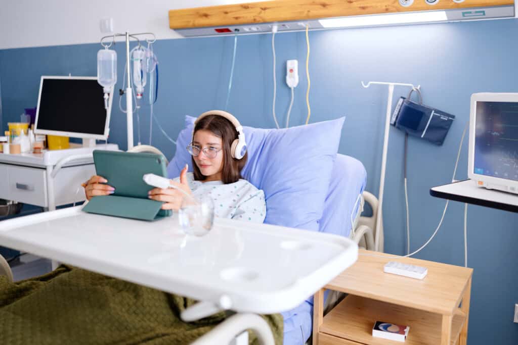 A patient in a hospital bed, possibly checking her hospital bag items on a tablet, wears headphones. She is wrapped in a green blanket amid medical equipment like a blood pressure monitor and IV stand. The room, with light blue walls and a bedside table, offers a calm setting.
