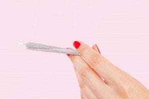 A hand with red-painted fingernails holds a rolled joint against a pink background, highlighting the controversial issue of marijuana use and pregnancy.