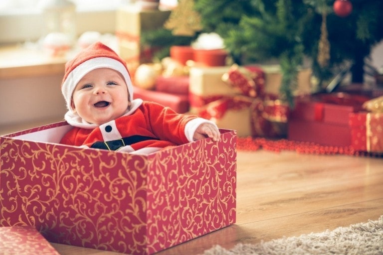 A baby dressed in a Santa outfit sits inside a red and gold patterned gift box, smiling while looking at the camera. In the background, there is a Christmas tree and several wrapped gifts on the floor, ready to prepare your kids to meet Santa.