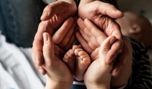 Two adult hands and one child's hands cradle the tiny feet of a baby, symbolizing new parent advice through the interconnected hands and feet. The baby is partially visible in the background, emphasizing care and togetherness in this tender moment.