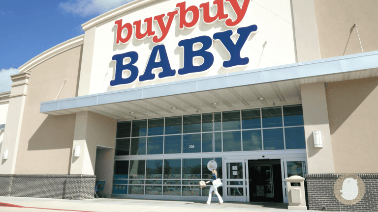 A person carrying a large stuffed animal is entering the famed buybuy BABY store. The exterior of the store features the name prominently at the top with large windows and automatic sliding doors.