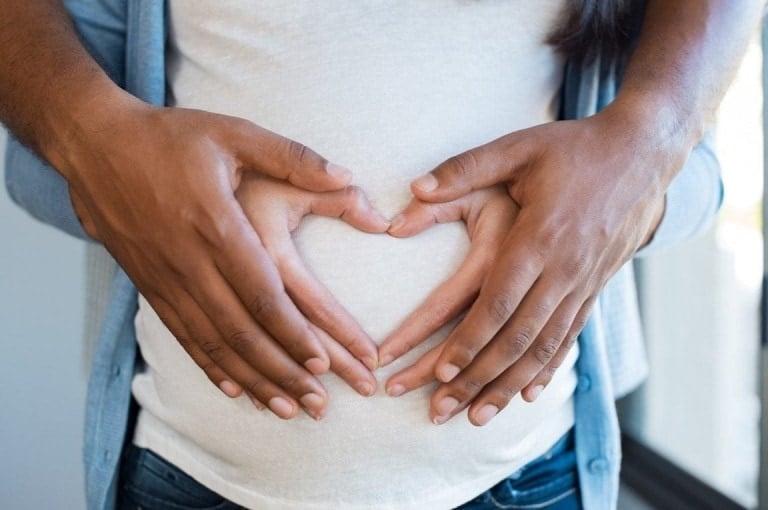 A close-up of two pairs of hands forming a heart shape over the belly of a pregnant person highlights major decisions pregnancy brings. The individual's hands are underneath, and the partner's hands are above, both creating the heart gesture. The person is wearing a white shirt and blue jeans.