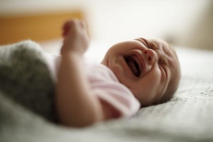 A fussy baby wearing a white outfit is lying on a soft surface, crying with eyes closed and mouth open. The background appears to be blurred, suggesting focus on the baby. One arm is raised, and a gray blanket partially covers the baby, common among breastfeeding babies needing comfort.