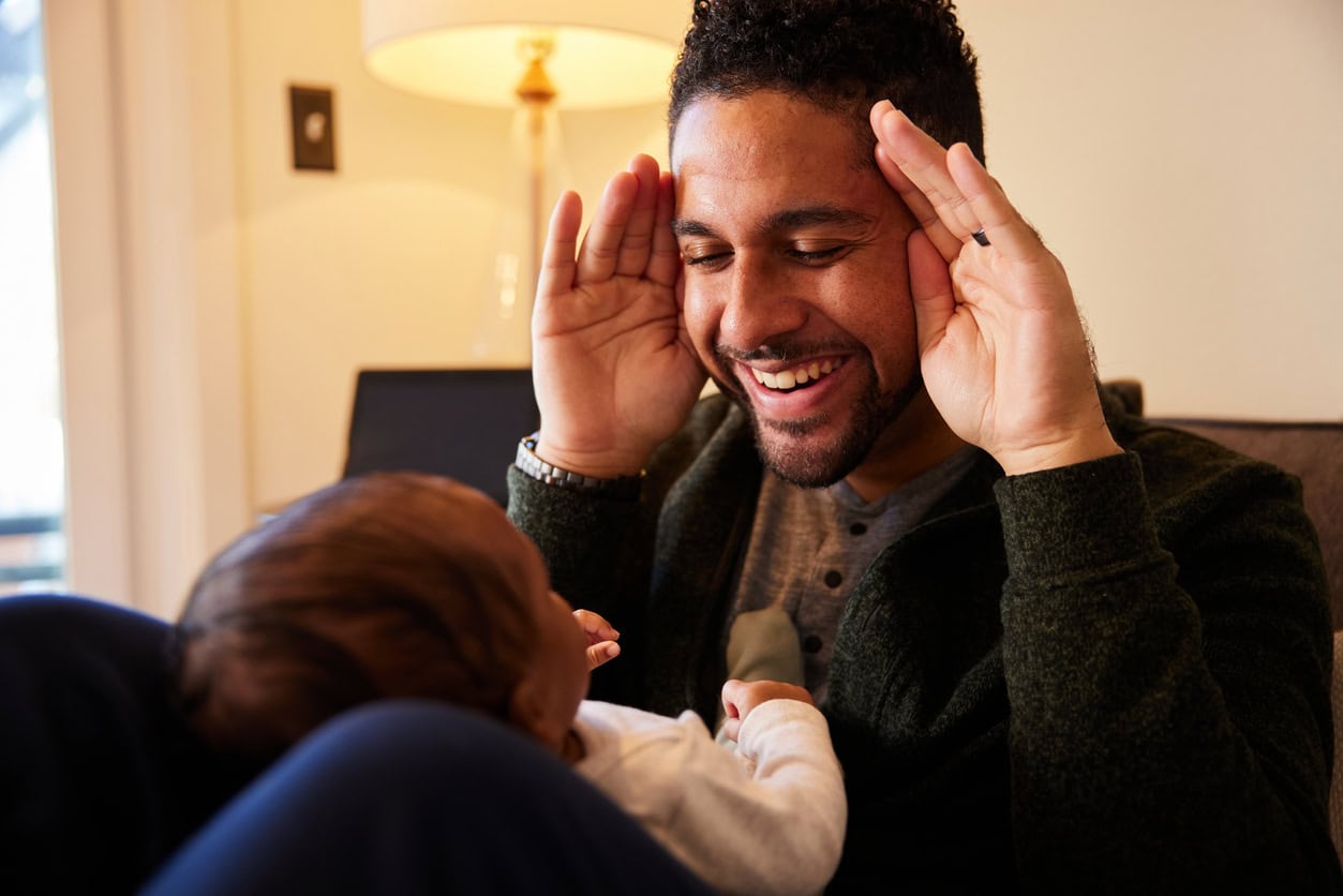 Smiling young father playing peekaboo with his adorable baby boy lying on his lap on a sofa at home