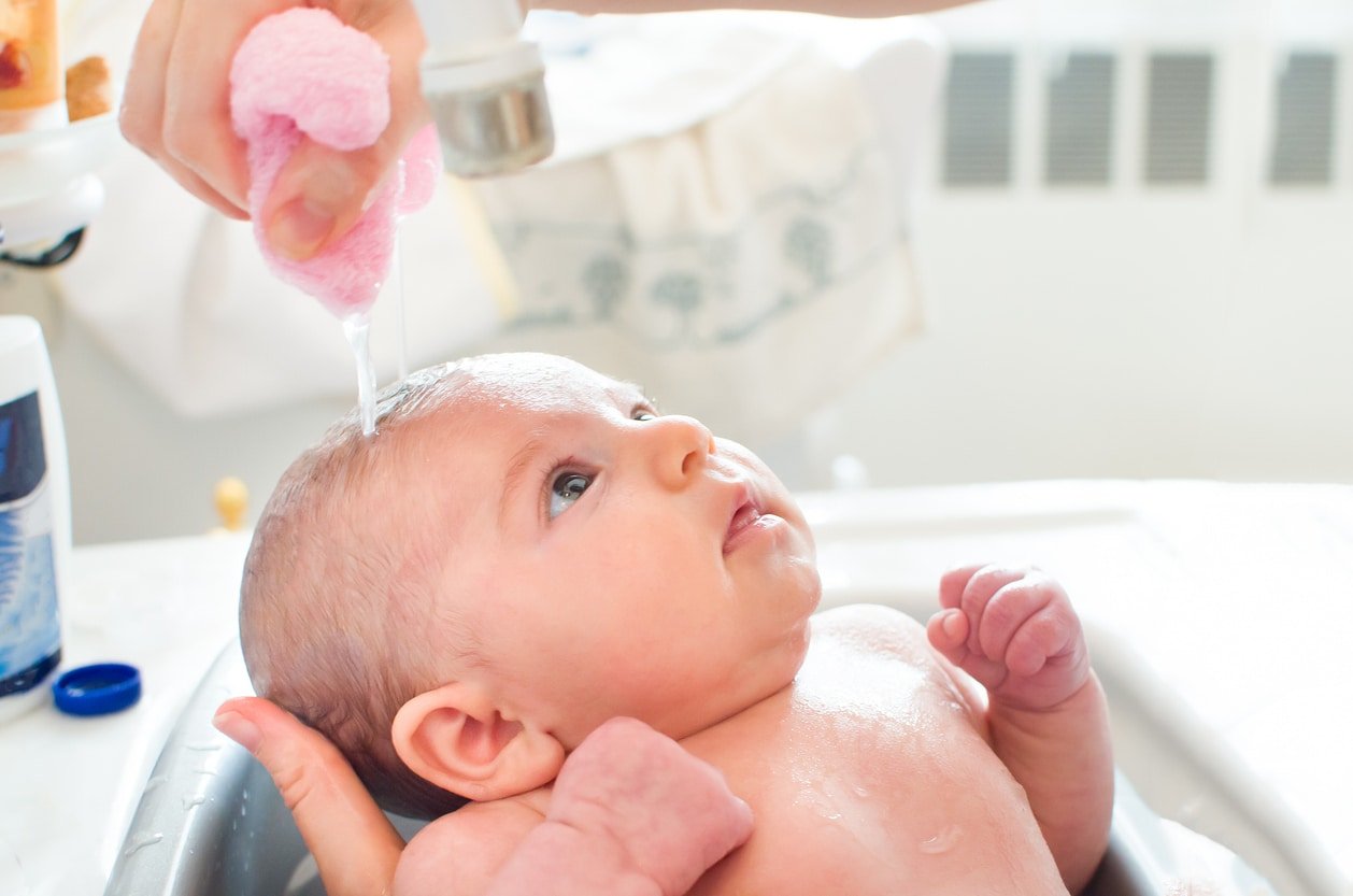 A newborn baby is getting a bath. Adult hands are holding the baby and pour water over baby's head with a washcloth.