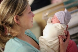 A woman holds a newborn baby wrapped in a white blanket with a striped hat, gazing lovingly at the sleeping infant while visitors at the hospital after birth gather in the brightly lit room.