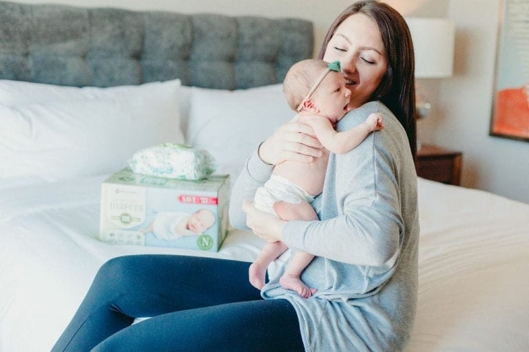 A woman is sitting on a bed, holding a baby who is wearing a diaper and a headband. Next to her, there's a box of Member's Mark diapers from Sam's Club and a stack of baby clothes. The woman is smiling and looking down at the baby.