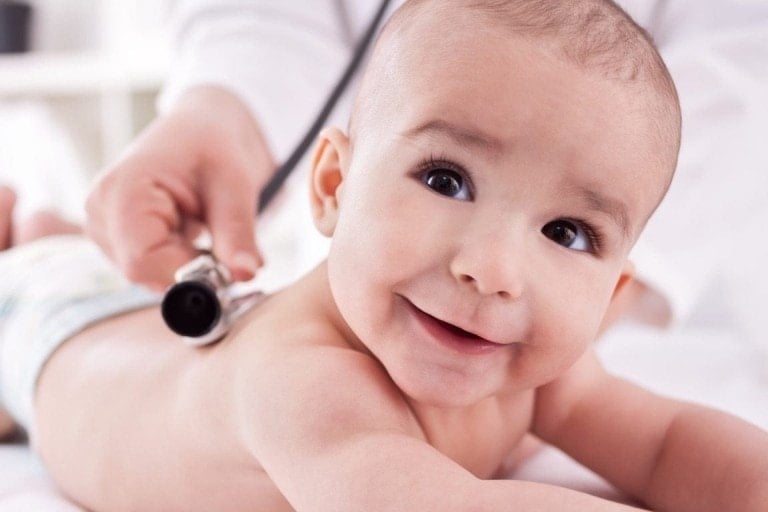 A baby lies on their stomach, smiling while a healthcare professional uses a stethoscope on the baby's back. The baby is wearing a diaper and appears relaxed, creating an ideal moment for interviewing pediatricians in this clinical setting.