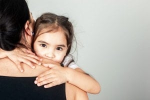 A young child with pigtails holds onto an adult with one arm wrapped around their neck. The child's face is turned towards the camera, while the adult has their back to the camera. The adult, clad in a black sleeveless top, appears to be offering comfort—a moment of time-in against a plain gray backdrop.