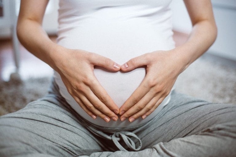 A person wearing a white tank top and gray pants sits cross-legged on the floor, forming a heart shape with their hands over their pregnant belly. This serene moment captures the calm before early labor, with focus on the torso and hands against a blurred background.