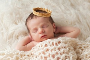 newborn baby laying on back covered in blanket wearing a small gold royal crown