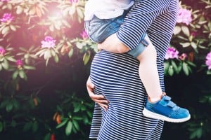 A pregnant person in a blue and white striped dress holds a small child wearing a matching striped shirt, denim shorts, and blue shoes. They stand in front of flowering bushes, perhaps already considering how to plan for their second baby.