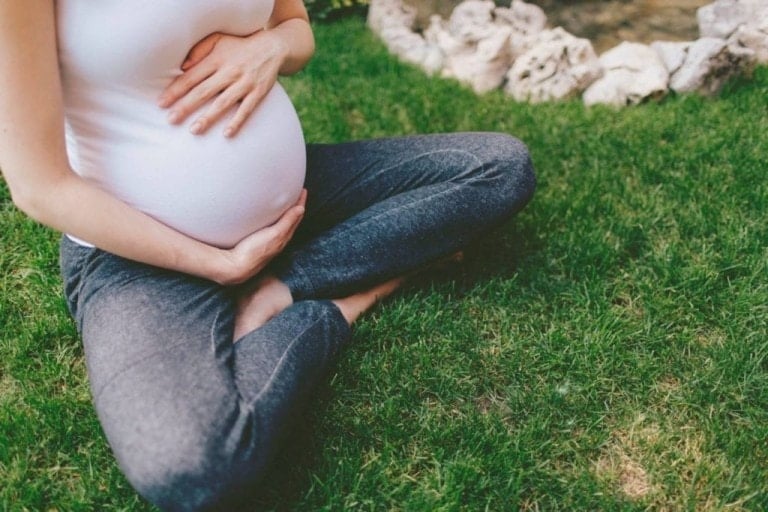 Pregnant woman sitting on grass, legs crossed, holding her pregnant belly.