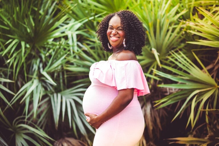 Beautiful glowing pregnant woman in pink dress poses for a maternity photo outdoors with palms in the background