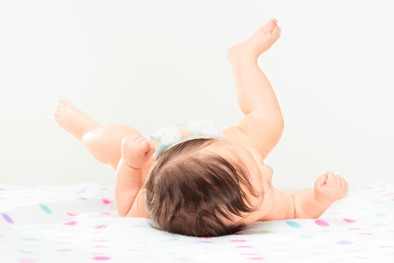 Back shot of little baby girl lying on polka dots blanket. Baby has got dark hair and legs raised above head. The background is bright and has plain color so it is easy to expand. The horizontal image.