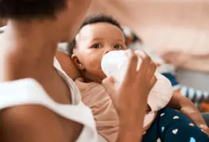 2 month old visit A mother holds her baby who is drinking from a bottle, wearing a light pink shirt and dark blue pants with white spots. The woman's face is not visible as she looks down, attentive to any baby full cues. The background is blurry with warm-toned furnishings.