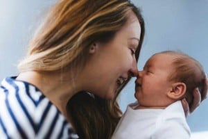 A woman with long hair lovingly touches noses with an infant she holds in her arms, capturing the pure joy of becoming a mom. Both are smiling, creating a tender and affectionate moment. The background is blurred and indistinct, with a blue hue.