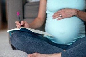 A pregnant person sits cross-legged on the floor, wearing a light blue tank top and gray leggings. They hold a book and a pen in their hands, one resting on their belly as they start to create a birth plan. The background features a sofa and a carpeted floor.