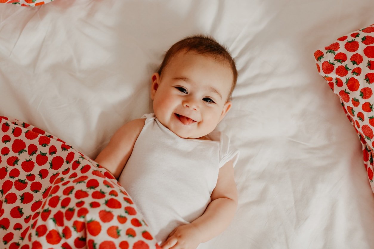 Happy baby girl laying down on a bed with a strawberry blanket smiling at the camera.
