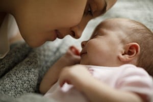 A close-up image of a person gently touching their nose to the nose of a sleeping baby. The baby, lying on a soft surface and wearing a light pink outfit, appears peaceful and content. The person is smiling warmly down at the baby, perhaps wondering when do babies first smile in response to such tender moments.