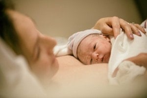 A newborn baby wrapped in a white blanket lies on its mother's chest as she gently cradles the baby's head with her hand. Wearing a striped hat, the baby looks awake and peaceful. The background is softly blurred, capturing the tender moment following the signs of labor.