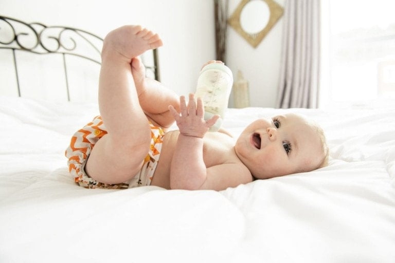 A Pretty baby girl drinks milk from bottle lying on bed. Child wearing diaper in nursery room.