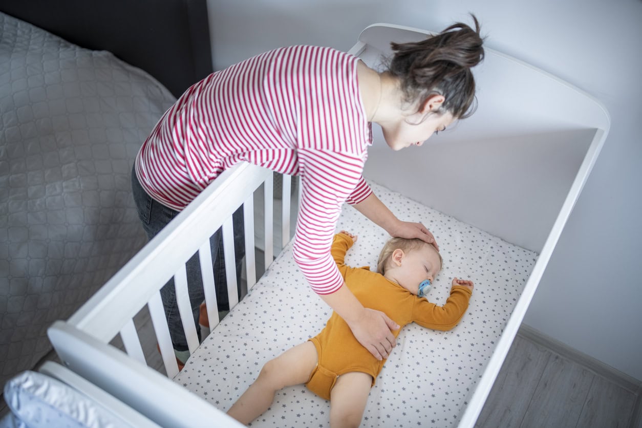 Caring young mother putting her baby boy for his nap in his crib.