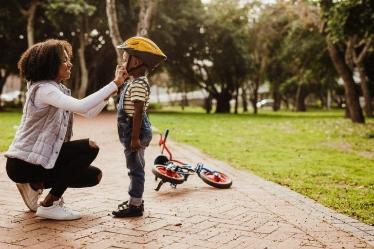 Mother helping son wearing helmet for cycling at park. Boy getting ready by wearing bike helmet to start cycling.