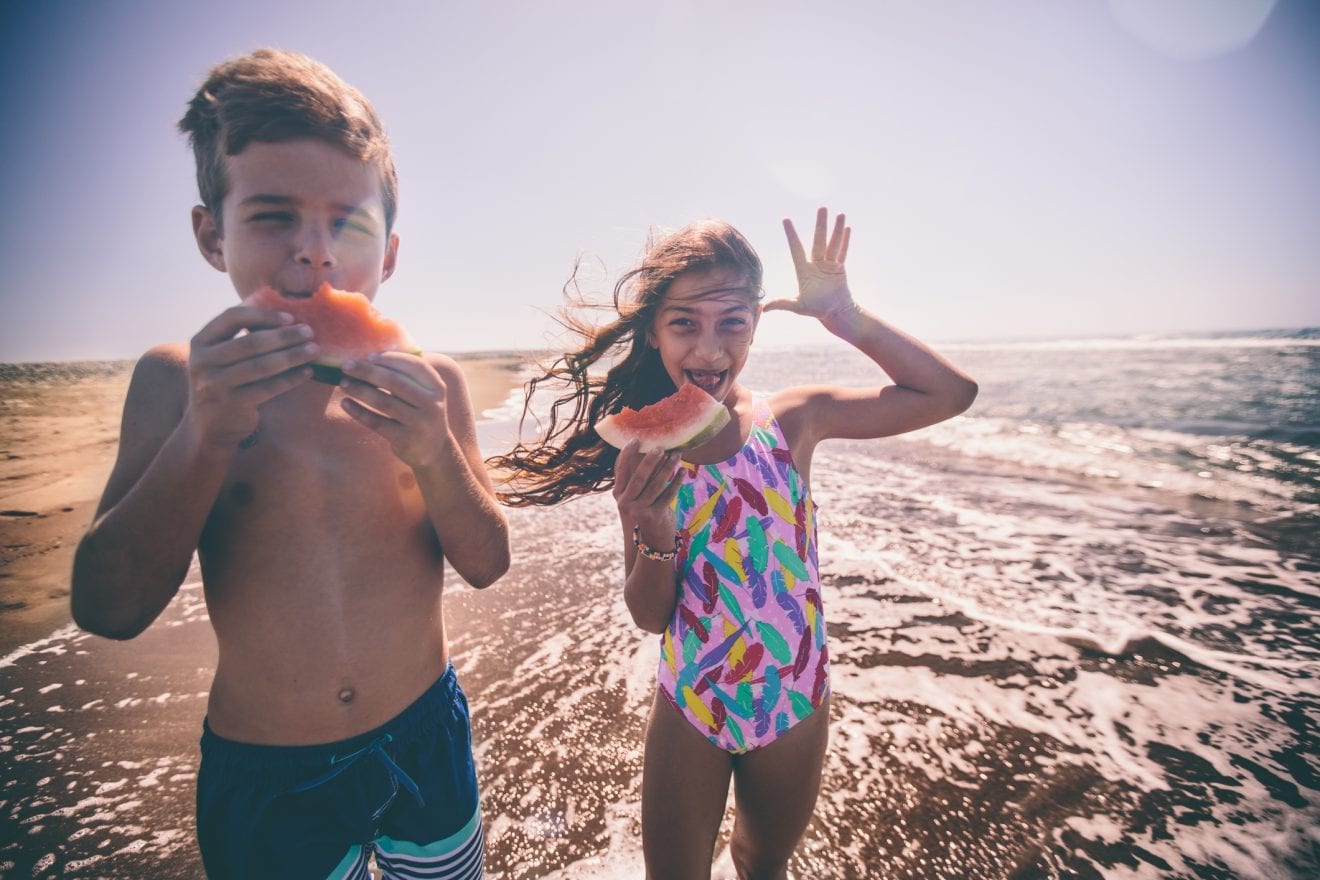 Brother and sister on family summer vacations making funny faces and eating watermelon at beach