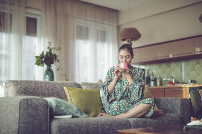 Young woman at home sitting on her couch drinking coffee in her robe.