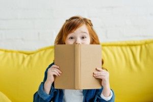 Child sitting on sofa and holding book in front of her face.