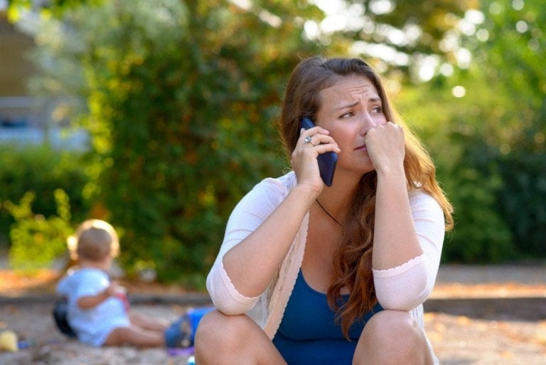 Stressed depressed young mother talking on a mobile phone as she sits outside in a playground with her baby son playing behind her.