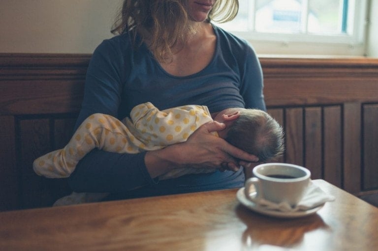 A young mother is breastfeeding her baby in a cafe while she is having a coffee.
