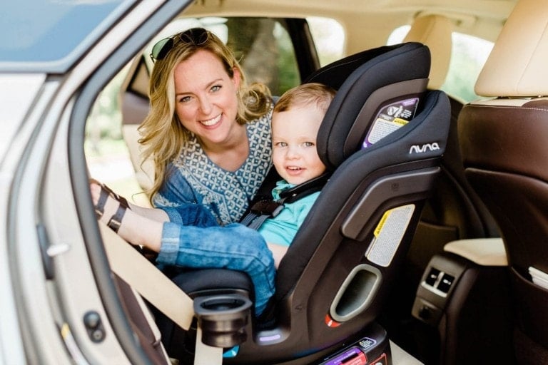 Toddler boy sitting in his all-in-one NUNA EXEC carseat with his mom helping him buckle in.