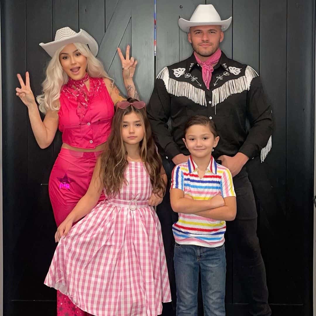 A group photo shows two adults and two children standing in front of a black wall. The adult woman, dressed in a pink outfit and white cowboy hat, makes peace signs. The adult man wears a black outfit and white cowboy hat. Celebrating in family Halloween costumes, the girl wears a pink checkered dress, and the boy sports a striped shirt.