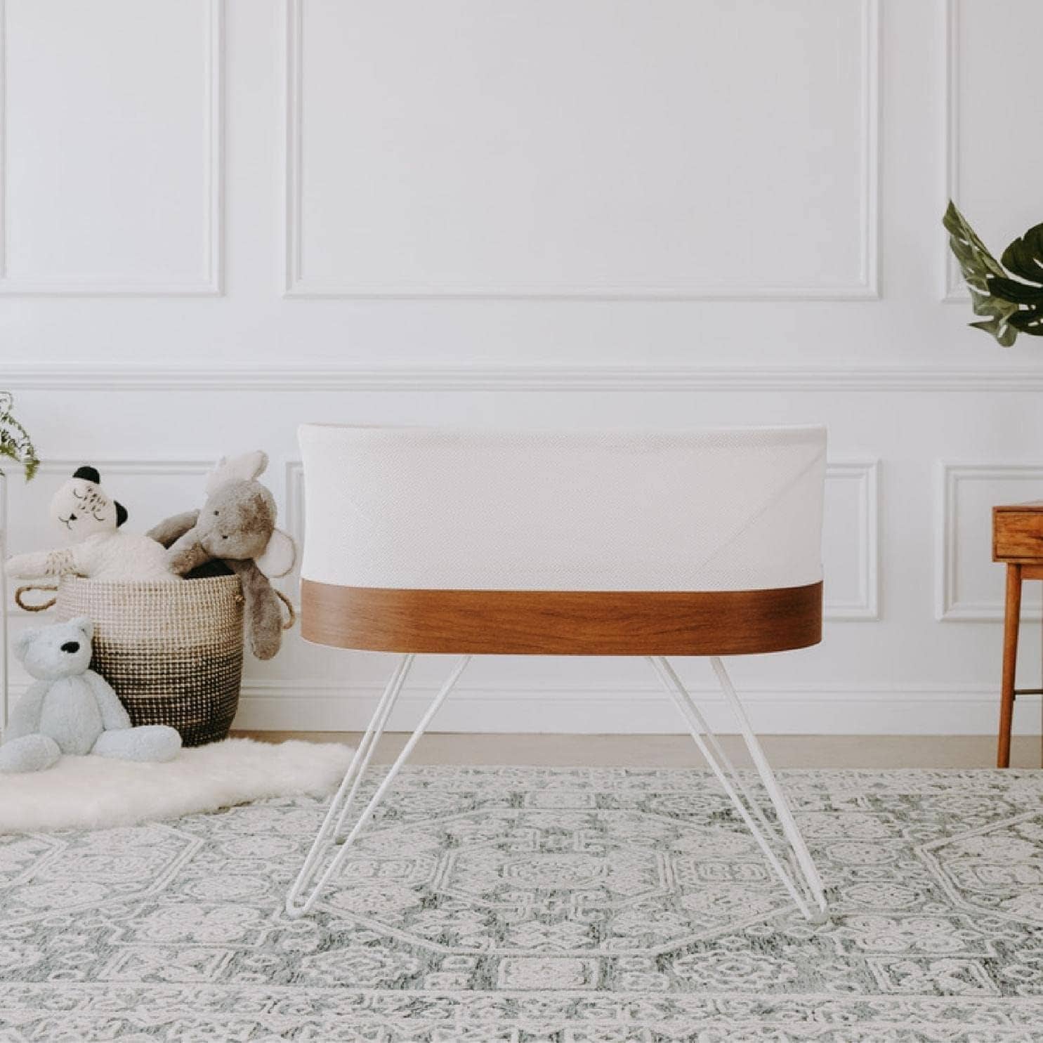 A modern white bassinet with wooden trim and thin metal legs stands on a patterned rug in a serene nursery. The room, perfect for postpartum recovery, boasts white paneled walls, a basket filled with stuffed animals, a small wooden table, and a potted plant on the side.