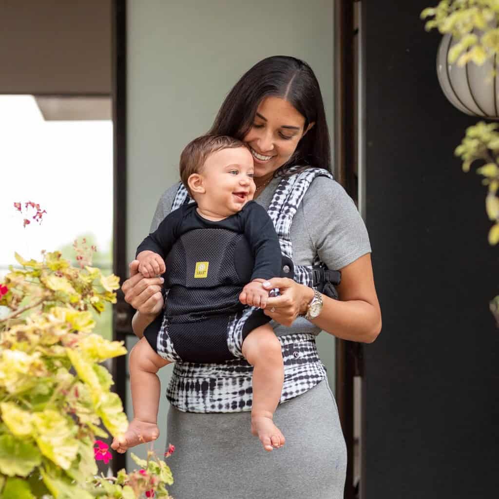 A woman standing outside near plants, smiling while holding a baby in a front-facing baby carrier. The baby, dressed in black clothing, is also smiling, and the woman wears a gray dress. Her radiant smile hints at the joy of successful postpartum recovery.
