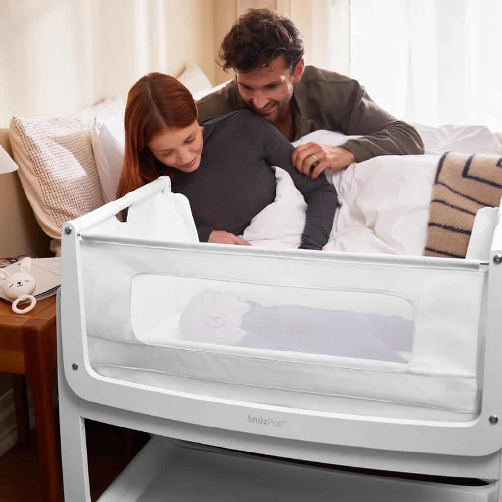 A couple lies in bed, looking at their baby asleep in a white crib attached to the side of the bed. The mother, focusing on postpartum recovery, has red hair and a gray shirt. The father has dark hair and a matching gray shirt. Their baby is swaddled in a white blanket, one of their postpartum essentials.