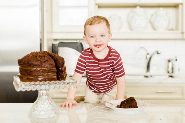 Toddler boy standing by a chocolate cake with some chocolate on his face.