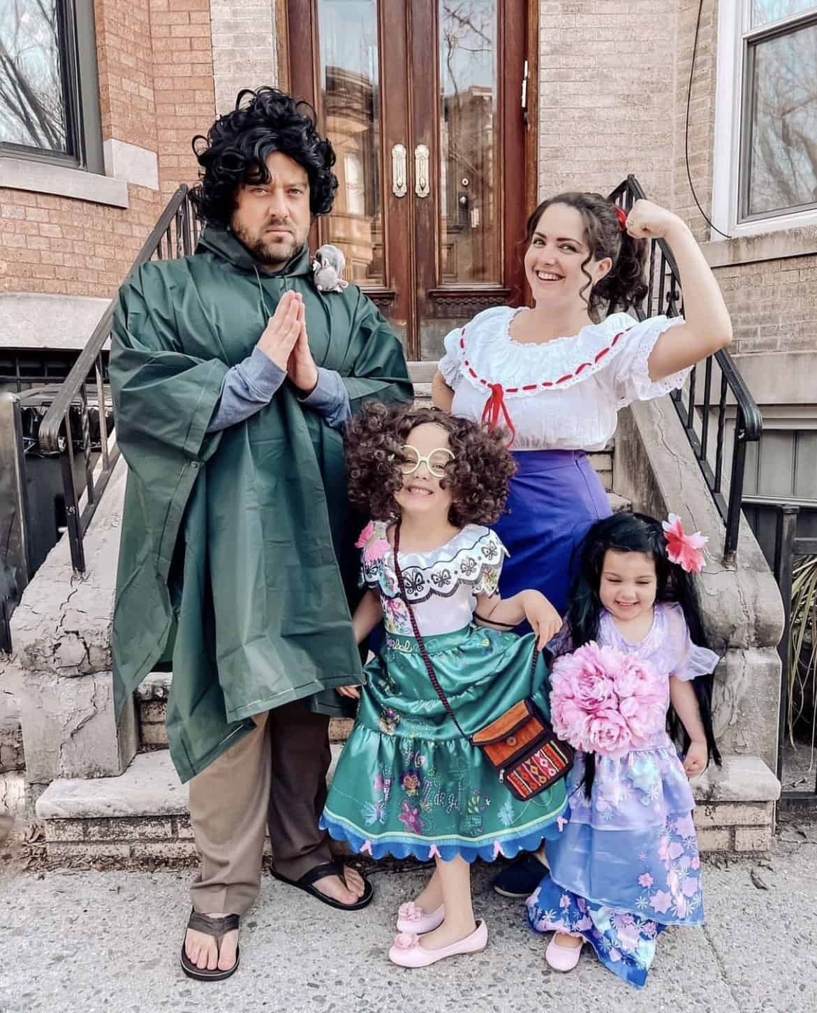A group of four people dressed in family Halloween costumes stands in front of a building. An adult man in a green cloak positions his hands in a prayer gesture. An adult woman in a white top and blue skirt flexes her arm, while two girls in colorful dresses pose cheerfully before them.