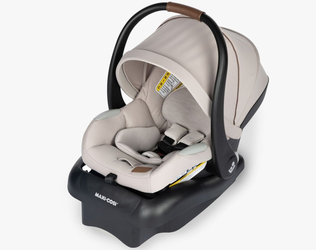 A beige infant car seat with a black base, featuring a brown handle and canopy—an essential for postpartum recovery. The car seat has a five-point harness and is labeled “MAXI-COSI” on the base, ensuring secure and safe transportation of infants in vehicles.