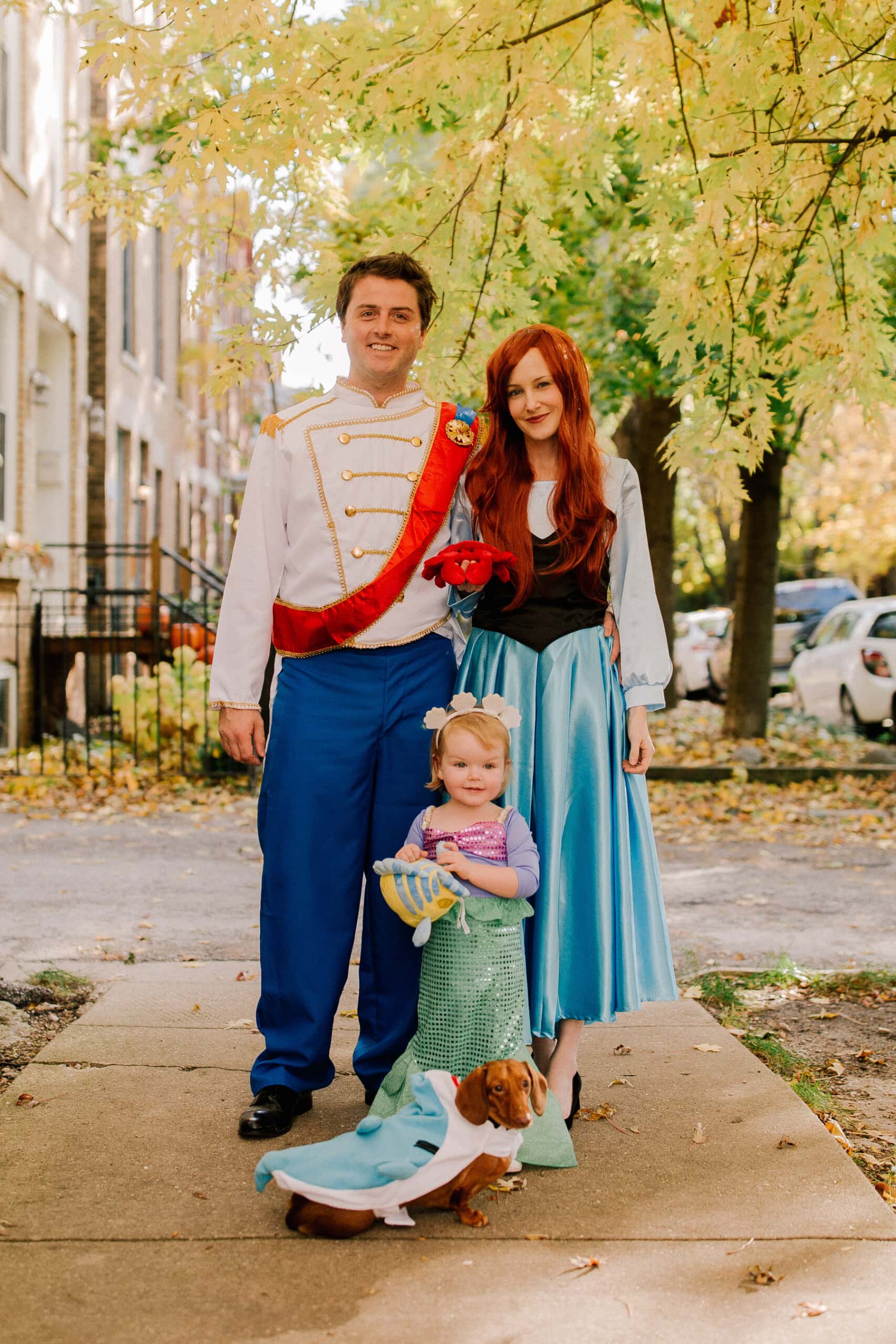 the little mermaid family costume, with dad dressed as prince eric, mom dressed as ariel in her land outfit, daughter dressed as ariel in her mermaid attire and holding flounder, and a dachshund dressed as a shark 