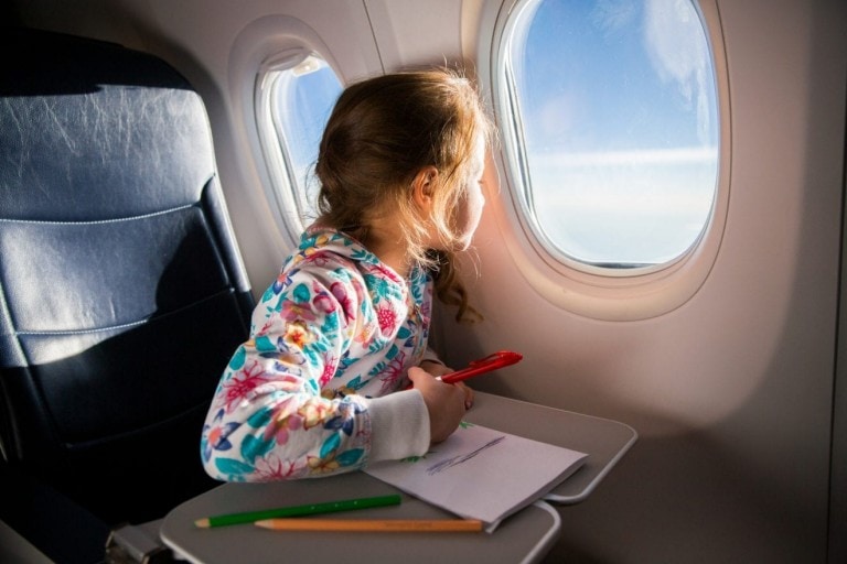 Child drawing picture with crayons in airplane. Little girl occupied while flying in aircraft. Travel with family and kids. Blue sky and sun outside the window.