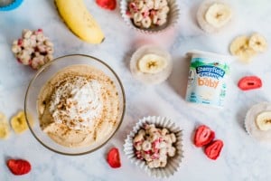 A bowl of whipped dessert topped with cinnamon, surrounded by banana, freeze-dried fruit, cereal clusters, and a container of Stonyfield Organic Baby yogurt on a marble surface.