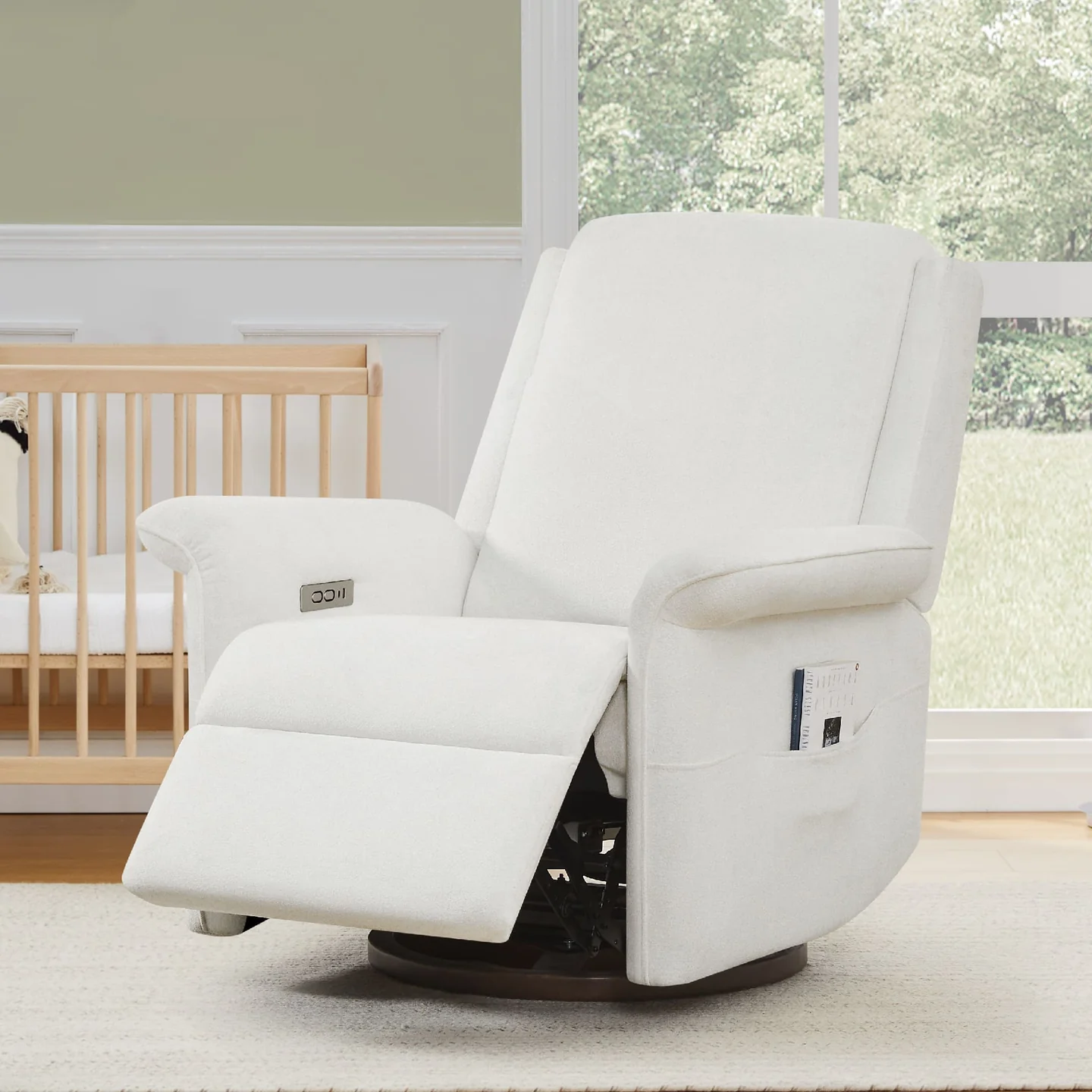 A white reclining chair, perfect for c-section recovery must-haves, with an extended footrest is positioned indoors near a window. It features a side pocket holding magazines and is situated next to a wooden crib. The room has light green walls and a beige carpet.