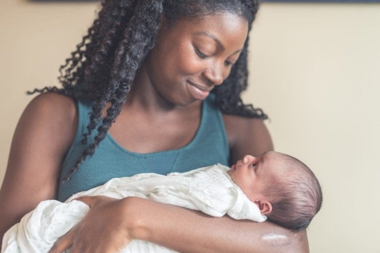 An African American mom gently cradles her newborn daughter in her arms. She is wrapped up and swaddled and is looking up at mom with a content expression. Mom tenderly gazing back at her.