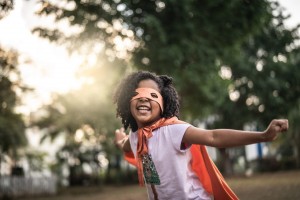 Smiling child wearing an orange superhero cape and mask stands outdoors with arms outstretched, with trees and sunlight in the background.